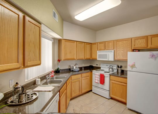 Modern kitchen here at Dobson Towne Centre Apartments with light wood cabinets, gray granite countertops, and white appliances. A red towel hangs on the oven, and two butterfly magnets adorn the fridge, creating a warm and inviting atmosphere.