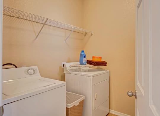 Compact laundry room here at Dobson Towne Centre Apartments with a white washer and dryer. Detergent and towels rest on the dryer. Beige walls and wooden floor create a clean, functional space.