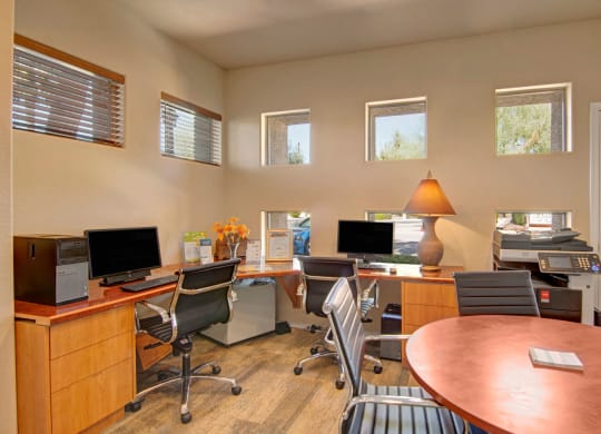 Modern office space here at Dobson Towne Centre Apartments with neutral tones, featuring two desks with computers, black chairs, a lamp, and a printer. Natural light streams through small windows.