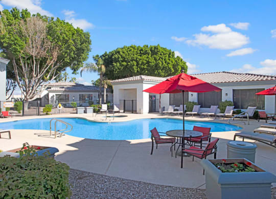 Sunny pool area here at Pala Mesa Apartments with turquoise water, surrounded by white lounge chairs. Red umbrellas provide shade. Trees and white buildings in the background. Relaxing vibe.