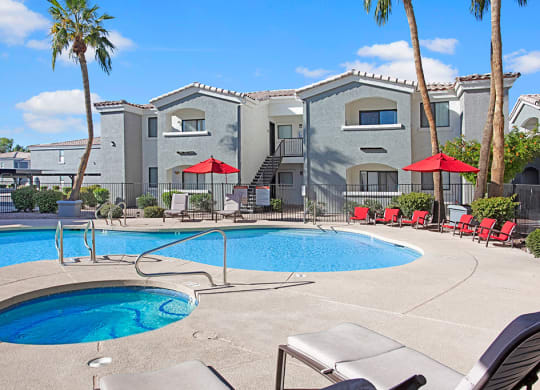 Pala Mesa Apartment complex with a pool and hot tub surrounded by lounge chairs and red umbrellas. Bright, sunny day with clear blue skies and palm trees.