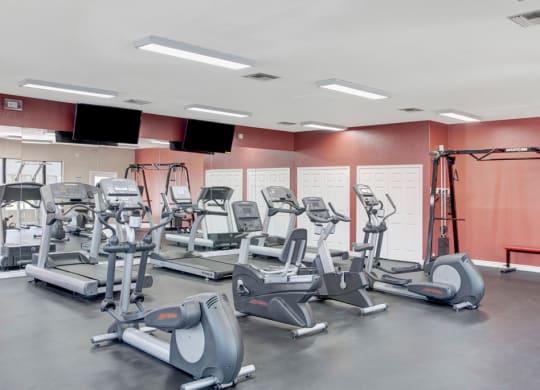 A modern gym here at Pala Mesa Apartments with various cardio machines, including treadmills and ellipticals, arranged on a dark floor. The room has white walls and two wall-mounted TVs.