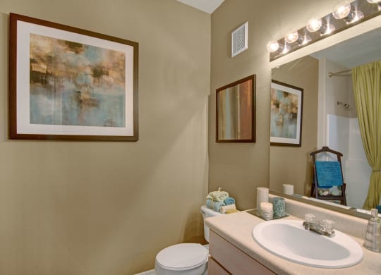 A cozy bathroom here at Pala Mesa Apartments with beige walls features abstract art, a wide mirror with bright lights, and neatly arranged towels. A shower curtain adds a touch of green.