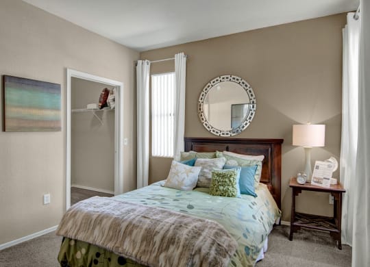 A cozy bedroom here at Pala Mesa Apartments with a wooden bed adorned with blue and green pillows. A circular mirror hangs above the headboard. Soft light from a table lamp on a wooden nightstand creates a warm ambiance. Light tan walls and white curtains enhance the serene atmosphere.