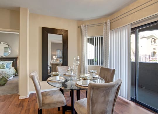 Dining area here at Pala Mesa Apartments with a glass table set for four, featuring beige velvet chairs. Large mirror and sliding doors brighten the space, leading to a cozy bedroom.