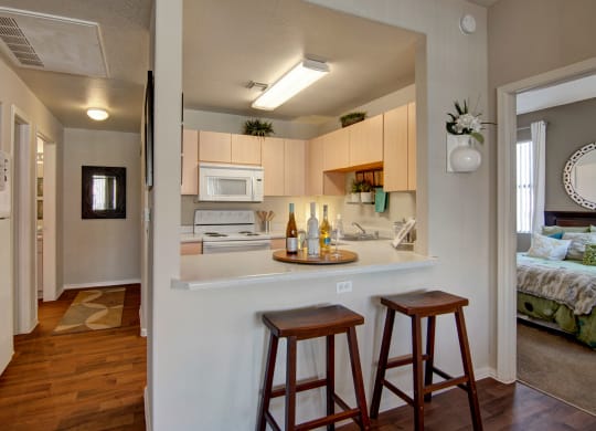 Compact kitchen here at Pala Mesa Apartments with light wood cabinets, a white stove, and microwave. Counter with wine bottles and glasses, two wooden stools. Cozy, inviting feel.