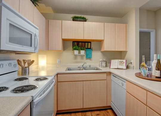 Modern kitchen here at Pala Mesa Apartments with light wood cabinets, beige countertops, and appliances including a microwave and stove. Decorative plants and wine bottles add warmth.