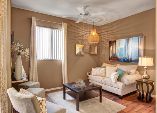 Cozy living room here at Pala Mesa Apartments with cream sofa, teal and patterned pillows, brown coffee table, floral vase, and lamp. Warm lighting and a serene ocean painting.