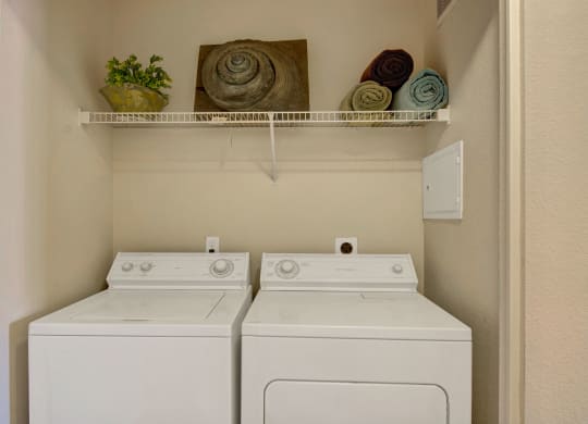 Compact laundry room here at Pala Mesa Apartments with a washer and dryer side by side. Above, a shelf holds a potted plant, three rolled towels, and spiral decor. Neutral tones.