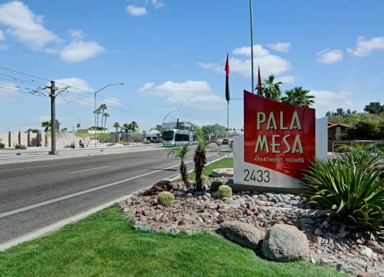 Landscape view of Pala Mesa Apartment Homes sign with palm trees and desert plants. A streetcar passes by on a sunny day, blue sky with clouds.