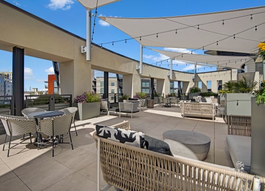 Rooftop terrace here at Roosevelt Row Apartments with modern seating, tables, and planters under white shade sails. String lights add ambiance. City skyline visible under a clear blue sky.