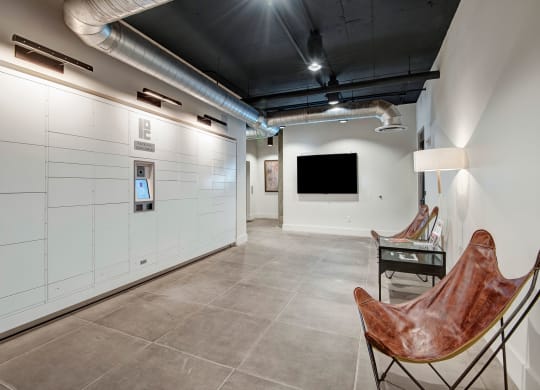 A modern lobby here at Roosevelt Row Apartments featuring white parcel lockers with a digital screen on the left. Two brown leather butterfly chairs and a lamp on the right, all under exposed ductwork.