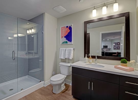 Modern bathroom here at Roosevelt Row Apartments with glass shower, dark wood vanity, and large mirror. Bright lighting, white towels, and art poster create a clean, stylish feel.