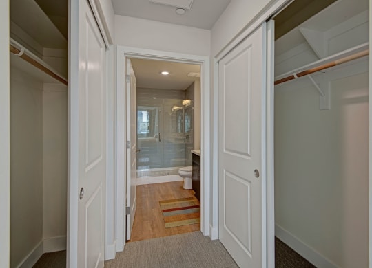 Hallway view here at Roosevelt Row Apartments of a white bathroom with clean lines and neutral tones. Dual closets flank the entry, leading to a glass shower and modern fixtures.