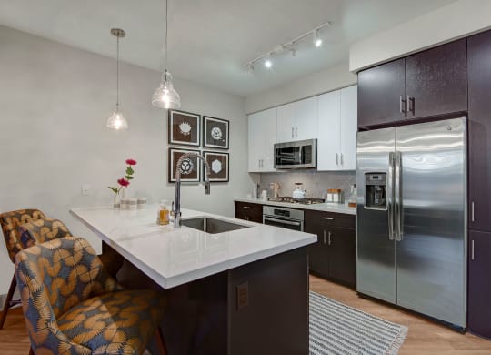 Modern kitchen here at Roosevelt Row Apartments with dark wood cabinets, white countertops, and stainless steel appliances. Two patterned chairs at the island, pendant lights, and floral decor add elegance.
