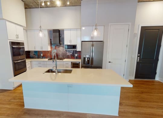 Modern kitchen here at Roosevelt Row Apartments with wooden floors, a large island with a sink, pendant lights, white cabinets, and colorful mosaic backsplash create a sleek look.