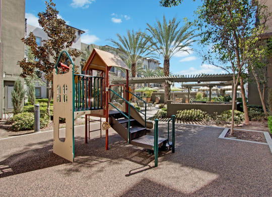 Playground here at Artisan at Main Street Metro Apartments with a small slide and steps in a sunlit courtyard, surrounded by lush greenery and palm trees, creating a peaceful and inviting atmosphere.