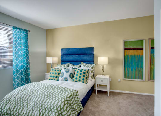 Contemporary bedroom here at Artisan at Main Street Metro Apartments with a blue upholstered headboard, patterned pillows, green comforter, and teal curtains. Abstract art adds vibrant color.