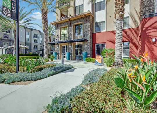 Exterior of our modern Artisan at Main Street Metro apartment complex with red accents, palm trees, and lush landscaping. A "Now Leasing" sign is displayed near the entrance.
