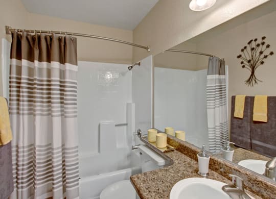 A clean, modern bathroom here at Castlepark Apartment Homes with neutral tones. Features a striped shower curtain, large mirror, yellow accents, and decorative wall art, creating a calming ambiance.