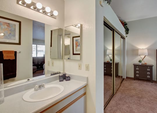Bright bathroom here at Castlepark Apartment Homes with a lit vanity mirror, geometric art, and towels. Adjacent bedroom has mirrored closet doors, a bedside lamp, and dresser. Cozy and inviting atmosphere.