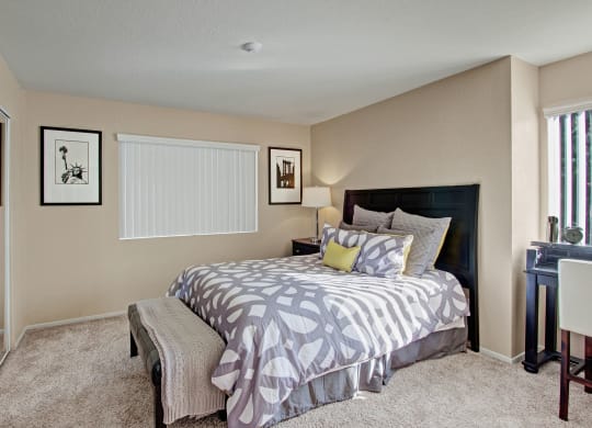 Cozy bedroom here at Castlepark Apartment Homes with a modern design. Features a bed with patterned gray and white bedding, a bedside lamp, abstract framed art, and a small desk by the window.