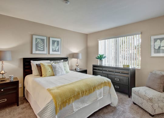 Contemporary bedroom here at Castlepark Apartment Homes with a double bed, decorative pillows, and a yellow throw blanket. Artwork adorns the walls, and a window lets in natural light.