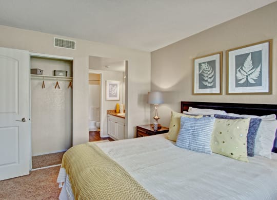 Cozy bedroom here at Castlepark Apartment Homes with a neatly made bed, yellow and gray pillows, fern artwork, open closet, and bathroom view. Warm, inviting ambiance.