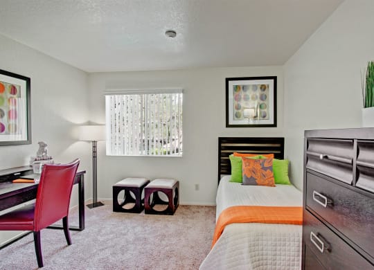 Cozy bedroom here at Castlepark Apartment Homes with a single bed featuring orange and green pillows. A desk with a red chair is on the left, and a tall dresser on the right. Soft lighting and abstract art enhance the modern feel.