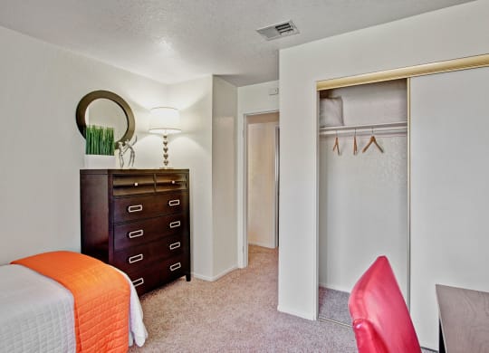 Bedroom here at Castlepark Apartment Homes with white walls, a bed with an orange cover, a tall dark dresser with a lamp and decor on top, an open closet, and a red chair next to a desk.