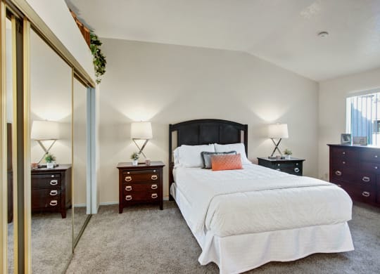 Cozy bedroom here at Castlepark Apartment Homes with a plush carpet features a central bed with white bedding, wooden headboard, two bedside tables with lamps, and mirrored closet doors.