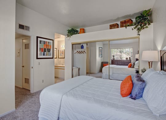 Bedroom here at Castlepark Apartment Homes with a cozy feel, showcasing a neatly made bed with white linens and orange accent pillows. Large mirror, decorative plants, and modern artwork adorn the walls. Soft lighting adds a warm ambiance.