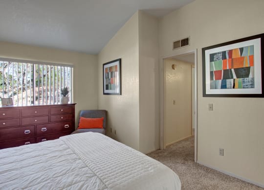 Bedroom here at Castlepark Apartment Homes with cream walls, featuring a bed with a white quilt, a dark wood dresser by a window, and colorful abstract wall art. Cozy and serene ambiance.