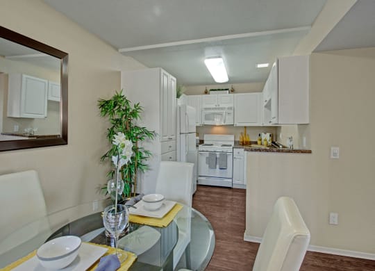 Modern kitchen and dining area here at Castlepark Apartment Homes with white cabinetry, a glass table, and leather chairs. A potted plant adds greenery; soft lighting creates a cozy atmosphere.