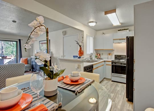 Modern kitchen and dining area here at Castlepark Apartment Homes with light wood flooring. A round glass table features white orchids, orange plates, and gray placemats, exuding a warm, inviting atmosphere.