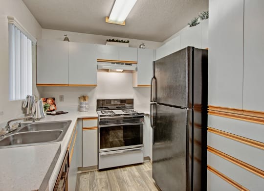 Compact kitchen here at Castlepark Apartment Homes with white cabinets, a black refrigerator, gas stove, and stainless steel sink. Bright fluorescent lighting and a tidy, modern appearance.
