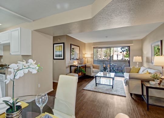 Bright, modern open-plan living room and kitchen here at Castlepark Apartment Homes. Features neutral tones, white cabinets, cozy sofas, large window with a view, and elegant decor.