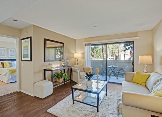 Chic living room here at Castlepark Apartment Homes with beige walls and dark wood floor. Cozy sofa with yellow pillows, glass table, mirror, and indoor plants. Sunny patio view through sliding doors.