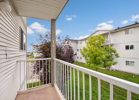 A cozy balcony here at Carrington Place Apartment Homes with white railings overlooks a grassy courtyard. Nearby, trees add greenery, while adjacent apartment buildings enhance the suburban feel.
