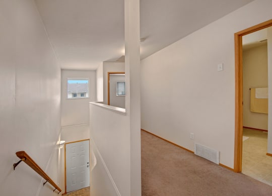 View of a simple, light-filled hallway here at Carrington Place Apartment Homes with white walls, a beige carpet, and a wooden railing on the stairs. A window with a view of rooftops is at the end, and a bathroom door with a towel is visible to the right.