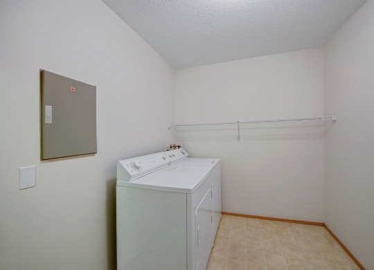 Laundry room here at Carrington Place Apartment Homes with white washer and dryer, beige tiled floor, white walls, and a wire shelf above the appliances. Clean, simple, and functional space.