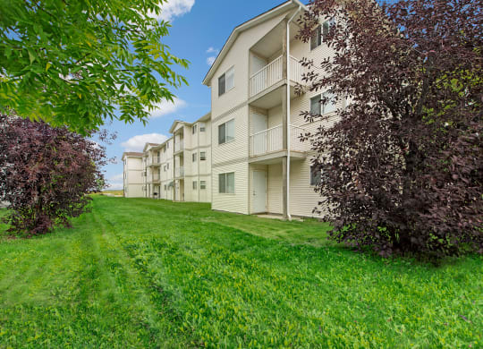 Carrington Place Apartment Homes buildings with beige siding line a lush, green lawn, bordered by trees with purple and green foliage against a blue sky with clouds.
