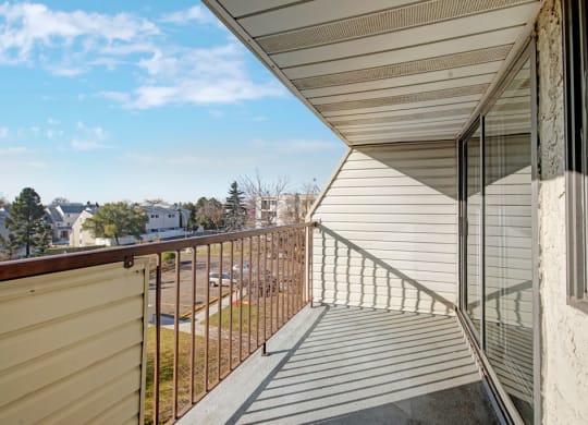 A sunlit balcony here at Cedar Gardens Apartment Homes with brown railings and sliding glass doors overlooks a scenic suburban neighborhood with trees and houses under a clear blue sky.