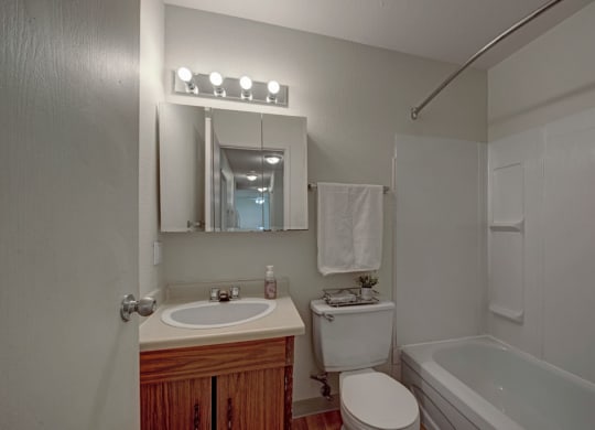 A small bathroom here at Cedar Gardens Apartment Homes with a wooden vanity, oval sink, and mirror cabinet. It includes a toilet, a white towel hanging, and a tub with a shower curtain rod. Bright lighting creates a clean and cozy atmosphere.