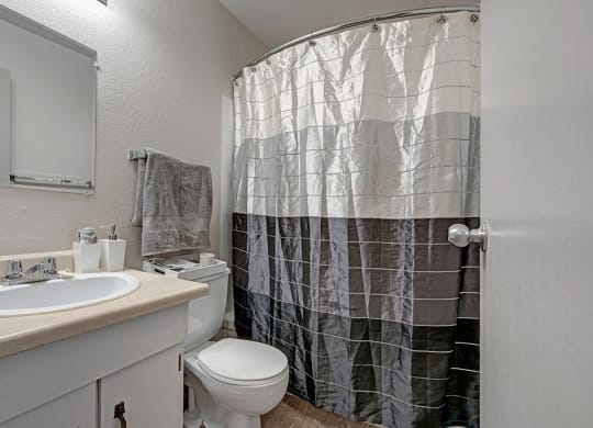 Small bathroom here at Cedar Gardens Apartment Homes with a beige countertop, white sink, and silver faucet. Gray towel hangs above the toilet. Black-and-white shower curtain adds contrast.