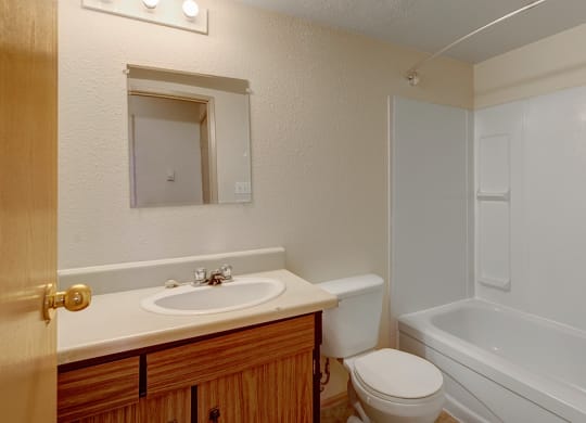 Small, clean bathroom here at Cedar Gardens Apartment Homes with beige walls. Features a wooden vanity, white sink, toilet, and bathtub with shower. Bright lighting above the mirror adds warmth.