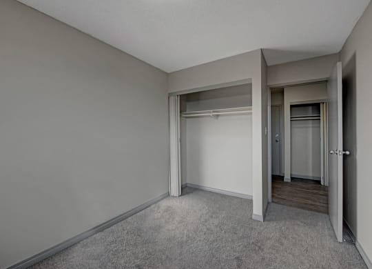 Empty bedroom here at Cedar Gardens Apartment Homes with gray walls and carpet, featuring an open closet with hanging rods. Doorway leads to hardwood floor hallway, creating a minimalist, neutral tone.