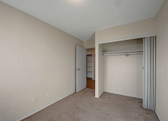 Empty beige room here at Cedar Gardens Apartment Homes with a carpeted floor, partially open closet with sliding door, and an open door leading to a hallway with hardwood flooring. Minimalist and neutral.