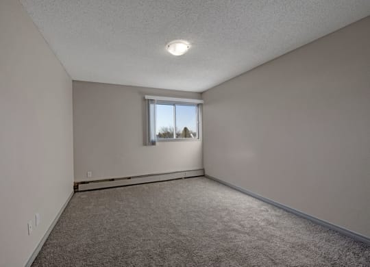 Empty bedroom here at Cedar Gardens Apartment Homes with beige walls, gray carpet, and a small window on the far wall with blinds. Ceiling light creates a soft, neutral atmosphere.