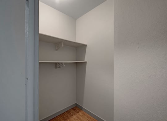 Small empty closet here at Cedar Gardens Apartment Homes with two simple white shelves on beige walls. The space is lit by a ceiling light, casting a neutral, minimalistic tone.